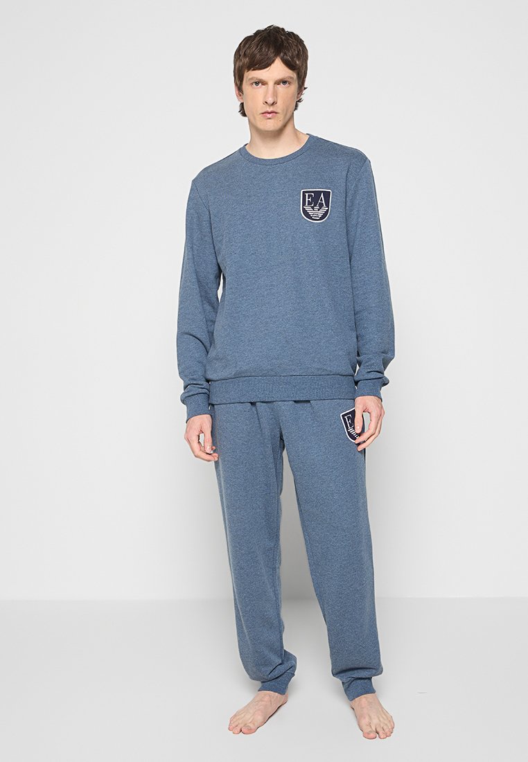 Homme portant un sweat-shirt à manches longues bleu et un pantalon de survêtement assorti avec des emblèmes EA, se tenant pieds nus devant un fond blanc uni.