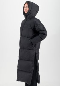 Manteau long noir rembourré avec une capuche, doté d'une fermeture éclair et de poches latérales. Le tissu a une texture lisse avec des motifs de couture visibles.