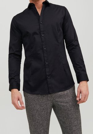 Man met een zwarte slim-fit overhemd met knopen en lange mouwen en grijze gestructureerde broek, staand tegen een effen lichte achtergrond.