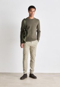 Olivno zeleno pleteno pulover, bež slim-fit hlače in temno rjave gležnjarje. Torba čez ramena v usklajeni zeleni barvi, gladka tekstura, minimalistična zasnova.