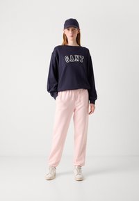 Námořnická mikina s nápisem "GANT", kombinovaná se světle růžovými tepláky, doplněná ladící čepicí a bílými teniskami. Jednoduchý design.