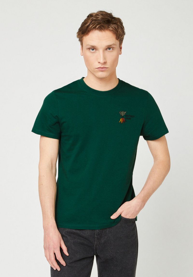 Jongeman met kort haar, gekleed in een donkergroen T-shirt met een klein kleurrijk groentedesign en tekst op de borst, donkere broek, hand in de zak.