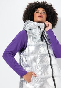 Silberne Puffer-Weste mit verstellbarem Kapuzenkragen, Reißverschluss vorne und zwei Seitentaschen, über einem lila Langarmshirt getragen.