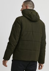 Man seen from the back wearing a dark green hooded puffer jacket with a patch on the upper sleeve, standing against a plain background.