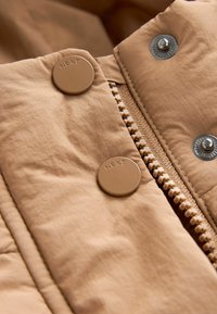 Gros plan sur une section de veste beige montrant des boutons-pression en plastique embossés du mot "NEXT" et une partie d'une fermeture éclair assortie avec des boutons-pression métalliques.