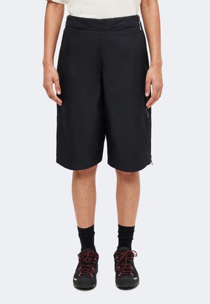 Personne portant un short noir jusqu'aux genoux, des chaussettes noires et des chaussures de randonnée noires avec des lacets rouges, debout devant un fond uni.