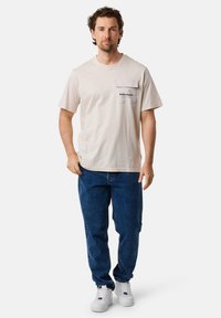 Beige bomull t-shirt med korta ärmar och en stor bröstficka med svart text. Bärs med blå denimjeans och vita sneakers.