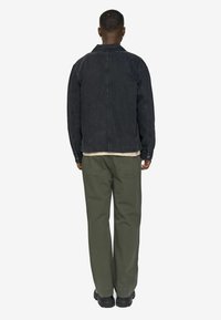 Man standing facing away wearing a black denim jacket, olive green pants, black shoes, and cream sweater peeking from under the jacket.