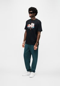 Μαύρο βαμβακερό μπλουζάκι με πολύχρωμο γραφικό και κείμενο, συνδυασμένο με πετρόλ φόρμες και λευκά αθλητικά παπούτσια. Το μοντέλο φορά γυαλιά ηλίου.