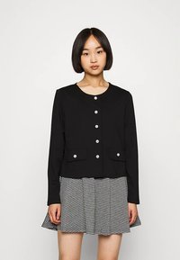 Giacca nera con bottoni davanti, due tasche, scollo rotondo e bottoni argentati lucidi, abbinata a una gonna a quadri nera e bianca.