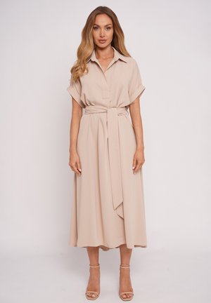 Kobieta o długich falowanych włosach, ubrana w beżową midi sukienkę z kołnierzykiem, z podwiniętymi rękawami, szerokim związanym paskiem oraz beżowymi sandałami na kostkę.