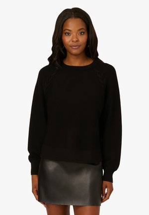 Maglione nero con dettagli intrecciati sulle spalle. Tessuto in maglia morbido e maniche lunghe, abbinato a una mini gonna in pelle nera lucida.
