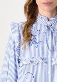 Stribet blå og hvid bluse med en flæsekrave, knaplukning og dekorativ blå broderi, samt særprægede mønstre på fronten.