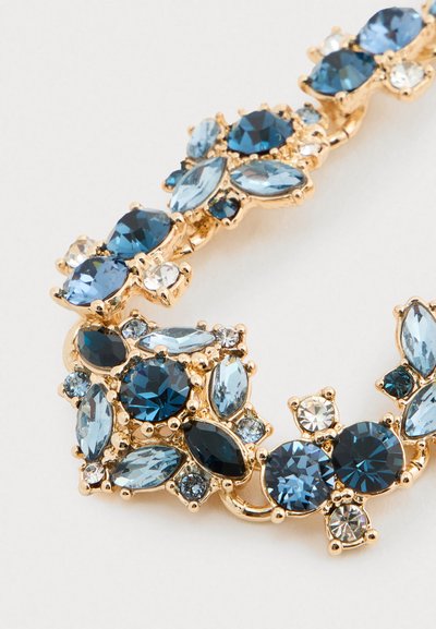 Gouden ketting segment met gegroepeerde ronde, ovale en marquise geslepen blauwe en heldere edelstenen op een lichte achtergrond.