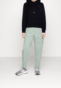 Czarna bluza z kapturem i sznurkami, jasne zielone spodnie cargo z bocznymi kieszeniami oraz szare sneakersy. Prosty design i luźny krój.