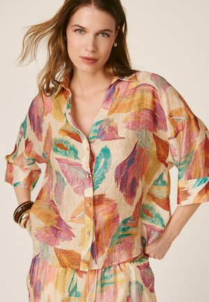 Joven mujer con camisa de botones y falda a juego con estampado abstracto de plumas coloridas, mangas remangadas y brazaletes en la muñeca.