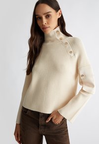Maglione a coste color crema con colletto alto, bottoni sulla spalla e maniche leggermente svasate, abbinato a pantaloni marroni.