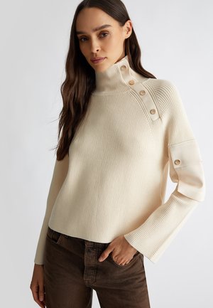 Maglione a coste color crema con colletto alto, bottoni sulla spalla e maniche leggermente svasate, abbinato a pantaloni marroni.