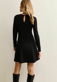 Vrouw met lang blond haar, gekleed in een zwarte jurk met lange mouwen en een open rug, en zwarte kousenlaarzen tot de knie, staand tegen een effen muur.