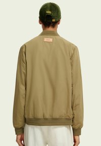 Beige Bomberjacke mit gerippten Bündchen und Saum. Markierung auf dem oberen Rücken. Kombiniert mit einer grünen Cordmütze. Glattes Material, lockere Passform.