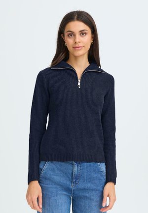 Marineblåt ribbet pullover-sweater med krave og halv lynlås. Har lange ærmer og et tætsiddende design. Bærer et par lyseblå jeans.