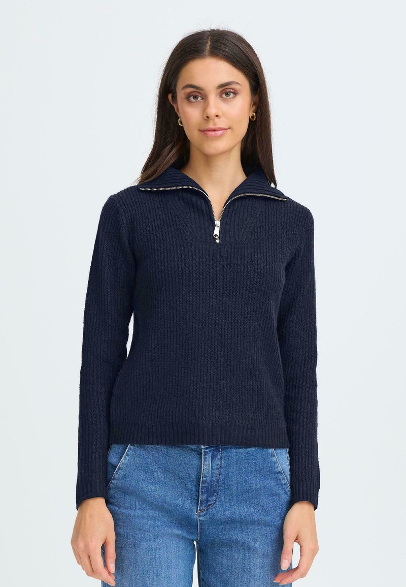 Maglione a coste navy con colletto e zip a metà. Presenta maniche lunghe e un design aderente. Indossato un paio di jeans azzurri.