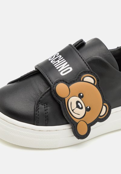 Sneaker in pelle nera con suola bianca, dotato di una applicazione a forma di orsetto e del branding "Moschino" sulla fascia. Texture liscia e design casual.