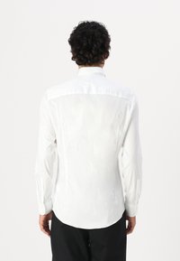 Camisa blanca de manga larga con una textura suave, cuello puntiagudo y puños abotonados. Ajuste entallado con un dobladillo curvado y un diseño minimalista.