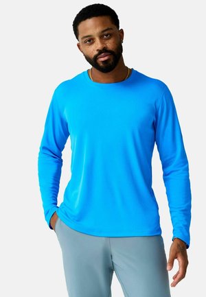 Uomo con barba e lentiggini che indossa una camicia blu brillante a maniche lunghe e pantaloni grigio chiaro, in piedi con una mano in tasca.