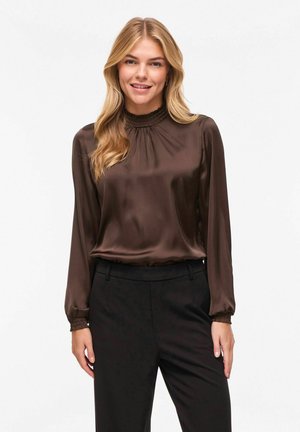 Femme blonde portant un chemisier en satin brun à manches longues avec col et poignets smockés, associé à un pantalon noir taille haute.