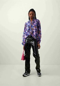 Fialová květinová košile na knoflíky s nabíranými rukávy, skombinovaná s černými lesklými kalhotami a růžovou kabelkou; bílá a černá obuv dodává vzhledu dokonalý finiš.