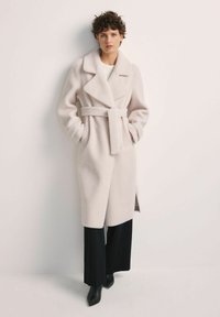 Kobieta w jasnobeżowym, wełnianym płaszczu z paskiem, nosząca czarne spodnie z szerokimi nogawkami i czarne botki na obcasie, stoi przy białej ścianie.