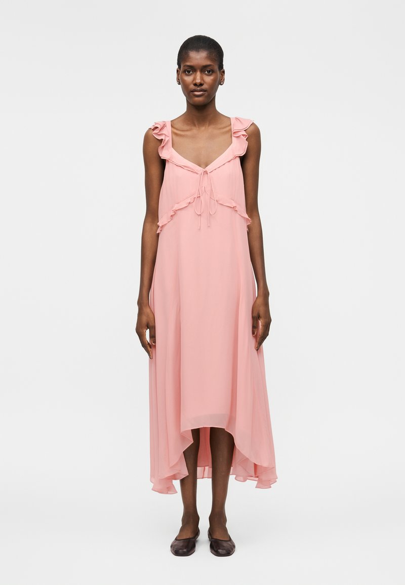 Γυναίκα που στέκεται φορώντας ένα αμάνικο, ανοιχτό ροζ μίντι φόρεμα με βολάν στους ιμάντες, δετό μπροστά και ασύμμετρο τελείωμα, σε συνδυασμό με σκούρα επίπεδα παπούτσια.