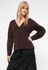 Καφέ πλεκτή ζακέτα με βαθύ V-λαιμόκοψη, που διαθέτει έξι κουμπιά μπροστά και μακριά μανίκια, σε συνδυασμό με μια υφασμάτινη μαύρη φούστα.