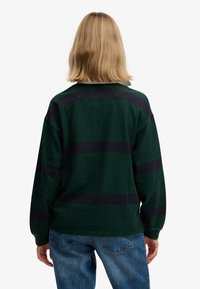 Personne aux cheveux blonds mi-longs, vue de dos, portant un pull vert foncé à rayures noires et un jean bleu.