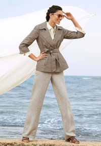 Lichtbruine op maat gemaakte blazer met strikdetail rond de taille, gecombineerd met losse beige broeken. Overgedimensioneerde zonnebril en bruine sandalen maken de look af.