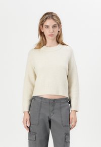 Maglione corto color crema con polsini e orlo a coste, abbinato a pantaloni cargo grigi con tasche laterali e dettagli di cucitura visibili.