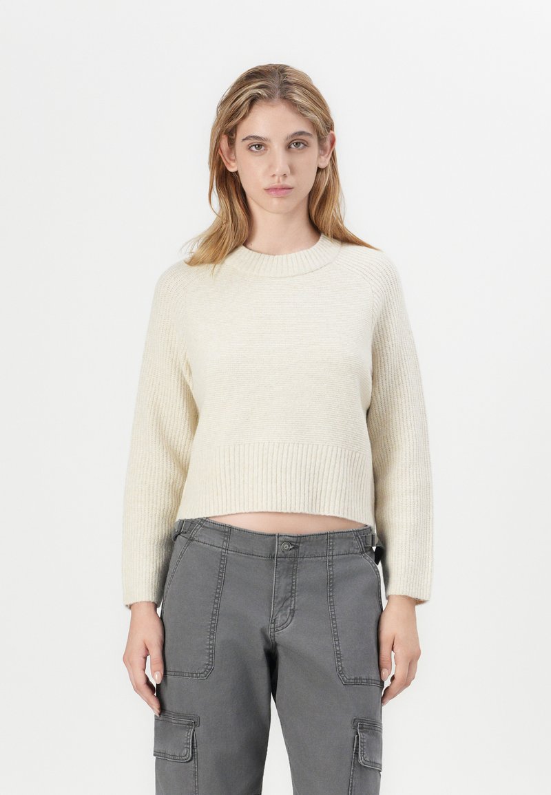 Maglione corto color crema con polsini e orlo a coste, abbinato a pantaloni cargo grigi con tasche laterali e dettagli di cucitura visibili.