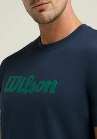Camiseta de algodón azul marino con un gran logotipo verde de "Wilson" impreso en el pecho. Diseño de mangas cortas y cuello redondo.