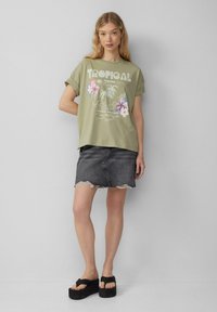 T-shirt surdimensionné vert olive avec un graphique "TROPICAL" et des accents floraux, associé à une jupe en denim noire et des sandales compensées.