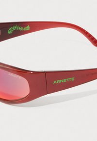 Crvene pravokutne sunčane naočale s roza lećama, zeleni logo "ARNETTE" sa strane i zeleni grafički prikaz soma u blizini odvojka okvira.