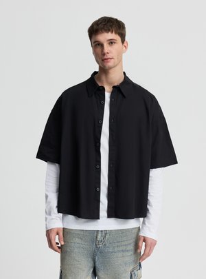 Giovane uomo che indossa una camicia nera a maniche corte sopra una camicia bianca a maniche lunghe e jeans azzurri, in piedi davanti a uno sfondo semplice.