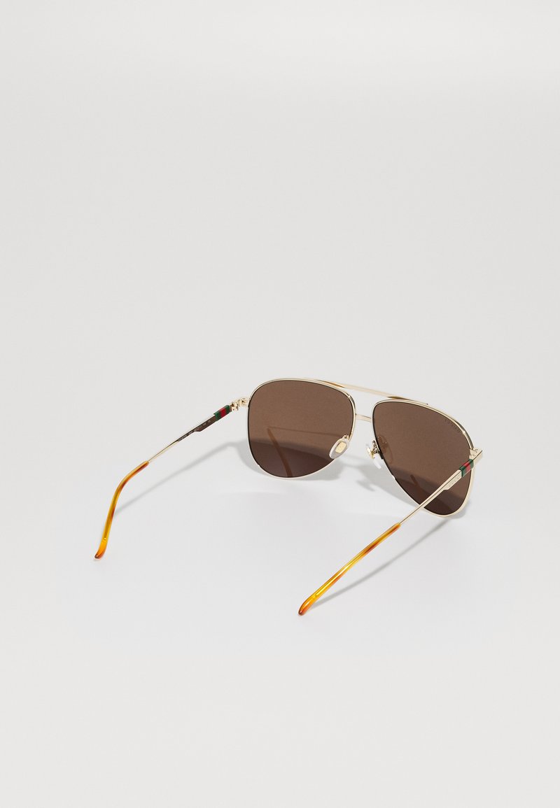 Occhiali da sole aviator con montatura in metallo dorato, lenti marroni scure e dettagli colorati a motivo sulle aste. Terminali auricolari in arancione e nero con texture.