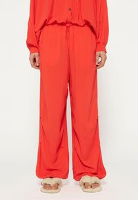 Rode wijde broek van lichtgewicht stof, met een elastische tailleband met trekkoord en zijzakken; gecombineerd met pluche slippers.