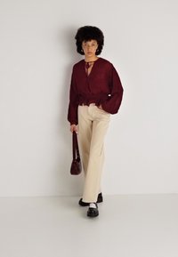 Blusa texturada em cor bordeaux com mangas soltas, decote amarrado; calças largas em creme; calçado escuro; pequena bolsa burgundy. Fundo neutro.