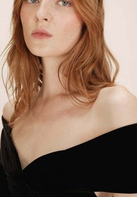 Femme aux cheveux roux avec des taches de rousseur portant un top noir à épaules dénudées, montrant son cou, ses épaules et une partie de son visage avec une expression neutre.