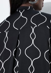 Primo piano di una persona che indossa una maglietta nera con motivi a linee ondulate bianche, mostrando la zona posteriore della spalla e parte del profilo.