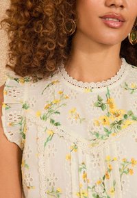 Blusa bianca con motivo floreale in giallo e verde. Presenta dettagli in pizzo ad occhio, maniche a cappello frill e un neckline ondulato.