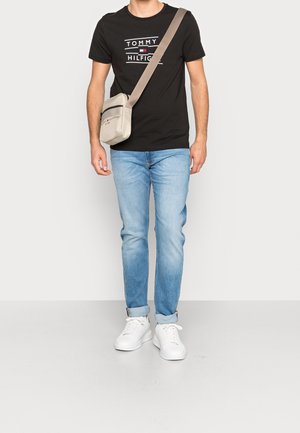 Zwart T-shirt met witte en rode afbeelding, lichtblauwe spijkerbroek, witte sneakers en een beige crossbodytas met ritssluiting en verstelbare riem.