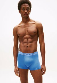 Giovane uomo con corporatura muscolosa che indossa boxer blu di Tommy Hilfiger, in piedi contro uno sfondo bianco.